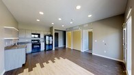 This single-floor space showcases a thoughtfully designed open-concept layout that seamlessly connects the kitchen and living areas. The kitchen is equipped with black appliances, including a refrigerator, range, and built-in microwave, complemented by light-toned cabinetry that provides ample storage. The countertops and cabinetry offer a clean, functional workspace suited for everyday use.

Durable hardwood-style flooring runs throughout the space, transitioning between two complementary tones that add visual interest and warmth to the interior. The recessed lighting installed across the ceiling ensures even, modern illumination throughout the open area.

The neutral gray walls and crisp white trim create a contemporary aesthetic that feels both clean and inviting. Ceiling vents indicate a centralized HVAC system for year-round climate control. Multiple doorways visible from the main living area lead to additional rooms, suggesting a well-connected floor plan with clear separation between living and private spaces. A dark accent wall near the entry door adds a bold design touch, grounding the space with contrast. Overall, the unit presents a modern, move-in-ready interior with practical finishes and a cohesive design sensibility.