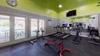 Located at 2930 SW 23rd St in Gainesville, FL, this fitness center is a well-equipped, single-floor workout facility designed to support a full range of exercise routines. The space is immediately distinguished by its bold lime-green upper walls and ceilings, which contrast sharply with the neutral gray lower walls, creating a vibrant and energetic atmosphere conducive to physical activity.

The flooring throughout is durable dark rubber matting, ideal for both cardio and weightlifting areas. Cardio equipment includes multiple treadmills and elliptical machines by TRUE Fitness, positioned along one wall and reflected in large wall-mounted mirrors that enhance the sense of space. A wall-mounted flat-screen television provides entertainment for cardio users.

The strength training section features a multi-function cable machine, a power tower for pull-ups and dips, a weight bench, and a dumbbell rack with free weights. A stability ball and additional accessories round out the functional training options.

Natural light floods the space through multiple sets of large French-style glass doors that open directly to an outdoor pool area, offering refreshing views and easy access to exterior amenities. A Coca-Cola vending machine is also visible near the pool entrance. Overhead pendant lighting complements the natural light, ensuring the space remains bright throughout the day.
