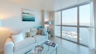 This thoughtfully designed single-floor residence offers a seamless blend of comfort and style, highlighted by breathtaking waterfront and marina views visible from both the living room and bedroom through expansive floor-to-ceiling windows. Natural light floods the open living area, which features light wood-tone flooring and a coastal-inspired palette of soft whites and aqua tones. The kitchen is outfitted with warm wood cabinetry, granite countertops, a stainless steel dishwasher, microwave, and a double sink, providing a fully functional cooking space. The bedroom is a tranquil retreat, with plush carpeting, generous natural light, and direct views of the marina, complemented by ample space for furnishings and a cozy seating area. The primary bathroom features a rich wood vanity with a white quartz countertop, large mirror, and a glass-enclosed walk-in shower with elegant tile surround. A separate half bath offers a pedestal sink and clean, neutral finishes. Throughout the residence, neutral walls, consistent tile and carpet flooring, and curated coastal artwork contribute to a cohesive, relaxing ambiance that is both refined and welcoming.