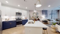 Located along Peachtree Road in Brookhaven, Georgia, this single-floor residence offers a thoughtfully designed interior that blends contemporary style with functional living. The open-concept kitchen is a standout feature, showcasing a bold two-tone cabinetry design with navy blue lower cabinets and crisp white upper cabinets, all accented with gold hardware. A large center island with a white quartz countertop and undermount sink serves as the focal point, complemented by elegant pendant lighting and stainless steel appliances including a gas range and French door refrigerator. Light wood-style flooring flows throughout, adding warmth and continuity. The living area is defined by a dramatic floor-to-ceiling abstract mural accent wall, creating an artistic backdrop for the bright, airy space. Natural light pours in through large windows dressed with sheer white curtains and blinds. The bedroom continues the navy blue accent wall theme, offering a cohesive design story, and features plush carpeting and natural light from multiple windows. The bathroom is finished with clean white tile, a glass-enclosed walk-in shower, white cabinetry, and wood-look flooring, delivering a crisp, modern aesthetic throughout.