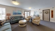 This single-floor community common area serves as a warm and inviting gathering space designed with residents' comfort and socialization in mind. The open layout is anchored by a generously sized lounge featuring multiple seating arrangements, including plush sofas, accent armchairs, and a tufted round ottoman, all coordinated in neutral and coastal-inspired tones that lend the space a calm, relaxed ambiance.

A standout feature of the space is a classic upright wooden piano, which serves as both a functional instrument and a charming focal point near the mailbox area. The resident mailbox wall and an adjacent counter provide practical amenities conveniently located within the communal space.

Natural light filters in through several windows and glass-panel doors, brightening the carpeted interior and complementing the soft, warm lighting provided by flush-mount ceiling fixtures. Coastal-themed artwork, including a nautical painting of sailboats on the water, adds a decorative touch that enhances the welcoming atmosphere.

The space also connects to a separate dining area, visible through an open passageway, which features traditional wooden chairs and tables. A reception or information window with decorative accents provides an additional point of engagement. Together, these elements create a cohesive, functional, and aesthetically pleasing environment suited for daily resident interaction and community life.
