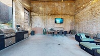 Located at 25 Draper St in Greenville, SC, this single-floor industrial space blends historic character with modern functionality. The open-plan layout is defined by striking floor-to-ceiling exposed brick walls that showcase the building's original craftsmanship, including a decorative arched brick fireplace alcove that serves as a dramatic focal point. Soaring ceilings amplify the sense of volume and openness throughout the space, while warm Edison-style string lights draped overhead create an inviting ambiance day or night. Large steel-framed warehouse windows flood the interior with abundant natural light and offer views of the surrounding greenery. The polished concrete floors complement the raw, industrial aesthetic seamlessly. The space is thoughtfully divided into distinct functional zones: a lounge area furnished with wicker sectional seating and teal cushions arranged around a coffee table, a dining area with a rustic wood communal table and chairs, and a wall-mounted flat-screen TV suitable for presentations or entertainment. An outdoor kitchen station along one wall features dual stainless steel grills and ample cabinetry, making the space well-equipped for catered events. A wide open-air entry arch connects the interior to the outdoor surroundings, enhancing airflow and accessibility. This versatile space is well-suited for gatherings, events, or creative activations.