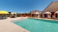 Located at 450 N Apache Rd in Buckeye, Arizona, this single-level outdoor amenity area is designed to deliver a resort-style experience in the heart of the Sonoran Desert. At its centerpiece is a generously sized rectangular swimming pool filled with crystal-clear turquoise water, accessible via stainless steel entry rails and equipped with in-floor fittings. The expansive pool deck is finished in a light aggregate surface that reflects the Arizona sun while remaining comfortable underfoot. Surrounding the pool, a well-appointed collection of yellow-framed lounge chairs and white-canopied umbrellas provides ample space for sunbathing and relaxation. A shaded gathering area with large cantilever umbrellas and vibrant red seating offers a social hub for residents and guests. The perimeter of the space features thoughtfully placed desert-adapted landscaping, including young trees and low-water shrubs set against decomposed granite, blending seamlessly with the arid environment. A corten steel fence lines the walkway at the front of the property, framing maturing trees and native plantings. The surrounding single-story buildings feature a mix of contemporary and traditional architectural elements, including gabled rooflines, stucco exteriors, and modern cladding. Under the vast, clear blue Arizona sky, this outdoor space offers a bright, open, and welcoming atmosphere year-round.