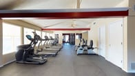 This single-story fitness center offers a spacious, open layout designed to accommodate a variety of workout routines. The room features vaulted ceilings with a prominent red-painted structural beam running across the length of the space, adding a bold architectural accent against the neutral cream-toned walls. A ceiling fan is mounted overhead, providing additional air circulation throughout the facility.

The flooring consists of durable dark rubber or foam interlocking tiles, well-suited for high-traffic exercise use. A row of treadmills and elliptical trainers lines the windowed wall, where multiple windows allow natural light to fill the space and provide exterior views during workouts. Recumbent bikes are positioned toward the center of the room, offering additional cardio options.

Toward the rear of the facility, strength training and cable equipment are visible, indicating a dedicated zone for resistance training. A mirrored wall in the background enhances the sense of depth within the space. A secondary doorway at the far end suggests possible access to an adjacent area or exit. The overall environment is clean, functional, and well-organized for comprehensive fitness use.