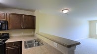 This single-story space in Fayetteville, North Carolina includes a kitchen area finished with dark espresso-toned cabinetry that provides a rich contrast against the light-colored walls. The countertops feature a speckled granite-style surface that extends along the perimeter, offering generous prep and workspace. A stainless steel double-basin sink with a standard faucet is integrated into the countertop, providing practical functionality. A peninsula or bar-style counter extension is visible, creating a natural divide between the kitchen and an adjacent area, which could serve as a dining or living space. The flooring transitions near this boundary, hinting at distinct zones within the layout. Flush-mount ceiling lighting provides overhead illumination, and standard electrical outlets are conveniently positioned along the countertop and walls. The walls are painted in a neutral, light yellow-green tone, contributing to a clean and understated aesthetic throughout. The overall design is straightforward and practical, offering a well-organized kitchen layout suited for everyday use in this single-level Fayetteville property.