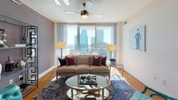 Located at 350 W 5th St in the heart of Uptown Charlotte, this single-floor residence delivers a compelling blend of contemporary design and urban convenience. Floor-to-ceiling windows are a defining architectural feature throughout the space, flooding each room with abundant natural light while framing striking panoramic views of the Charlotte skyline.

The living area is generously proportioned, featuring warm hardwood flooring, a ceiling fan, and sheer drapery that softens the expansive glass walls. A decorative open shelving unit adds visual interest alongside the neutral, muted wall tones. The kitchen is well-appointed with rich wood cabinetry, granite countertops, a subway tile backsplash, stainless steel appliances including a refrigerator, range, microwave, and dishwasher, and track lighting overhead.

The primary bedroom benefits from the same dramatic window line, offering direct skyline views, plush carpeting, and an en-suite bathroom complete with a soaking tub, separate glass-enclosed shower, and tile flooring. A secondary bedroom features an accent wall in deep olive, ceiling fan, and carpeted floors. A second full bathroom includes a frameless glass shower enclosure with clean tile finishes. Together, these spaces create a polished, upscale urban living environment.