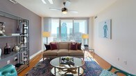 Located at 350 W 5th St in the heart of Uptown Charlotte, this single-floor residence delivers a compelling blend of contemporary design and urban convenience. Floor-to-ceiling windows are a defining architectural feature throughout the space, flooding each room with abundant natural light while framing striking panoramic views of the Charlotte skyline.

The living area is generously proportioned, featuring warm hardwood flooring, a ceiling fan, and sheer drapery that softens the expansive glass walls. A decorative open shelving unit adds visual interest alongside the neutral, muted wall tones. The kitchen is well-appointed with rich wood cabinetry, granite countertops, a subway tile backsplash, stainless steel appliances including a refrigerator, range, microwave, and dishwasher, and track lighting overhead.

The primary bedroom benefits from the same dramatic window line, offering direct skyline views, plush carpeting, and an en-suite bathroom complete with a soaking tub, separate glass-enclosed shower, and tile flooring. A secondary bedroom features an accent wall in deep olive, ceiling fan, and carpeted floors. A second full bathroom includes a frameless glass shower enclosure with clean tile finishes. Together, these spaces create a polished, upscale urban living environment.