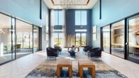 This single-story lobby presents a striking first impression through its double-height ceilings, which amplify the sense of openness and grandeur throughout the space. The interior is defined by a rich blue color palette across the walls, complemented by crisp white structural columns and light-toned large-format tile flooring laid in an elegant herringbone pattern.

Floor-to-ceiling windows line multiple walls, flooding the space with natural light and offering views of the surrounding exterior landscape. Sheer floor-length drapery frames the windows, adding softness to the otherwise bold architectural elements. A sculptural driftwood installation serves as a focal point against the rear wall, introducing an organic, nature-inspired contrast to the contemporary design.

The seating area is thoughtfully arranged with dark upholstered lounge chairs, a velvet blue sofa, sleek round coffee tables, and ribbed leather ottomans atop a large abstract area rug with blue and gold tones. An oversized fiddle-leaf fig in a ceramic planter adds a lush, living element near the entry. Textured wall art in neutral tones is displayed throughout, while a contemporary chandelier overhead ties the space together with a refined, modern ambiance.