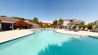 Located at 8108 Vinoy Blvd in Charlotte, North Carolina, this outdoor amenity space offers a resort-inspired experience designed for relaxation and outdoor entertaining. At its centerpiece is a generously sized, freeform swimming pool with crystal-clear turquoise water, surrounded by a wide, smooth concrete sundeck. The deck is furnished with lounge chairs and shaded umbrella tables, providing ample space for both sunbathing and casual dining poolside.

Adjacent to the pool is a well-appointed pergola structure anchored by stacked stone columns and topped with a white-painted open-beam roof strung with globe lights, lending warmth during evening gatherings. Beneath the pergola sits a fully equipped outdoor kitchen featuring a stainless steel gas grill, a compact refrigerator, and dark granite countertops set into a stone-faced base — ideal for outdoor cooking and entertaining.

The entire area is enclosed by a wrought iron fence with stone pillar accents, offering both security and visual cohesion with the surrounding architecture. Mature trees and manicured landscaping frame the space, providing natural shade and a lush backdrop. Under the clear Carolina skies, this outdoor amenity area delivers an inviting, community-focused environment suited for year-round enjoyment.