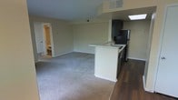 This single-floor space is designed with an open-concept layout that seamlessly connects the kitchen and living areas. The kitchen is outfitted with dark espresso-toned cabinetry, a stainless steel refrigerator, and additional appliances, all anchored by a white peninsula that doubles as a functional workspace or casual dining counter. The peninsula also houses a sink, adding to the kitchen's practicality. The kitchen features hardwood-style flooring, while the adjoining living area transitions to plush wall-to-wall carpeting in a neutral beige tone, creating a comfortable and cohesive aesthetic throughout. The walls are finished in a warm off-white, contributing to a bright and airy atmosphere. A skylight above the kitchen area introduces natural light, enhancing the overall sense of openness. Recessed HVAC vents are integrated into the ceiling for a clean, unobtrusive look. Toward the back of the living space, an open doorway leads to a bathroom, adding convenience and accessibility. The overall design balances functional living with a straightforward, clean aesthetic suited for everyday use.