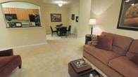This single-floor space showcases an open-concept layout that integrates the living, dining, and kitchen areas into one cohesive environment. The living area features plush, neutral-toned carpeting that extends throughout, creating a warm and comfortable atmosphere. A cushioned sofa accented with decorative pillows anchors the space, complemented by a small side table with a classic lamp that adds ambient lighting.

The dining area is situated adjacent to the living space and accommodates a dark-wood table with matching chairs, offering an intimate setting for meals. Framed artwork on the warm beige walls adds a decorative touch, contributing to the overall welcoming ambiance. Crown molding along the ceiling provides an elegant architectural detail that elevates the interior's finish.

A pass-through window connects the dining area to the kitchen, offering a glimpse of wood-toned cabinetry and countertop space beyond. This functional feature enhances the flow between spaces while maintaining a sense of openness. The ceiling-mounted light fixture provides overhead illumination, working in tandem with the floor lamp to create a layered, well-lit environment throughout.