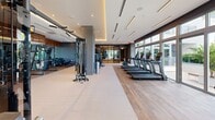 Located at 9600 S Dixie Hwy in Miami, FL, this state-of-the-art fitness center offers a comprehensive and elegantly designed workout environment on a single floor. The space is defined by its modern aesthetic, combining warm wood-tone flooring, cool grey accent walls, and a coffered ceiling with ambient lighting that creates a focused yet inviting atmosphere.

The facility is divided into distinct workout zones. A dedicated cardio area lines the perimeter, featuring a row of Matrix treadmills and elliptical machines positioned directly in front of expansive floor-to-ceiling windows. These large glass panels flood the space with natural light and frame lush tropical landscaping, offering an energizing outdoor connection while exercising.

The strength training zone is equipped with a full array of Matrix cable machines, adjustable benches, and a well-stocked dumbbell rack, providing versatile options for resistance training. A functional training area includes medicine balls, kettle bells, and a dual cable station, catering to a wide range of fitness routines.

A glass-walled studio space is accessible via sleek black-framed sliding doors, offering a semi-private area suitable for stretching, group classes, or personal training sessions. Mirrored walls throughout enhance the sense of openness. The overall design balances high-end finishes with practical functionality, making this a premium fitness destination in Miami.