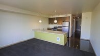 This single-floor residence offers a thoughtfully designed open-concept layout that seamlessly connects the kitchen and living areas. The kitchen is a standout feature, anchored by a spacious island with a light stone countertop and a vibrant lime-green base that adds a bold, modern touch. The island includes an integrated sink and provides ample workspace, doubling as a casual dining or entertaining surface. Overhead, elegant pendant lights hang at varying heights, casting a warm, focused glow over the island and enhancing the contemporary aesthetic. The kitchen is well-equipped with stainless steel appliances, including a refrigerator, range, and microwave, complemented by rich dark wood cabinetry that extends to the upper walls, offering generous storage. Track lighting installed along the ceiling ensures the workspace is well-lit throughout. The adjoining living area features plush dark carpeting that contrasts nicely with the smooth, polished flooring in the kitchen, creating a clear yet cohesive distinction between zones. Neutral-toned walls throughout maintain a clean, airy feel. A hallway visible from the main space hints at additional rooms beyond, rounding out this well-appointed and functional single-level home.