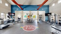 This TSC fitness center is a spacious, single-floor gym designed to support a full range of workout routines. The space is immediately striking thanks to its soaring vaulted ceilings accented by bold red exposed beams, set against vibrant teal-blue upper walls. Natural light floods the interior through a series of clerestory windows positioned high along the walls, creating an open and energizing atmosphere.

Distinctive natural stone columns divide the main floor into functional workout zones while adding a refined architectural character. The rubberized flooring throughout is both durable and practical, and features the TSC logo prominently at the center of the space.

The cardio zone is well-appointed with multiple treadmills, elliptical trainers, stair climbers, stationary bikes, and a rowing machine — all positioned near large windows that offer views of the surrounding outdoor area. A wall of mirrors amplifies the sense of space in this section.

The strength training area includes a multi-functional cable machine, Atlantis equipment, a comprehensive free weight rack, kettlebells in varying sizes, medicine balls, resistance bands, battle ropes, and foam rollers, all neatly organized on dedicated storage racks.

A colorful mural depicting cyclists adds a dynamic visual focal point to the rear wall. The facility is completed by dark wood-framed glass entry doors and a vending machine, making for a fully self-contained fitness experience.