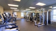This fitness center offers a spacious, single-floor layout purpose-built for effective and comfortable workouts. The room features a durable dark rubber flooring ideal for heavy equipment and high-traffic use, while a drop-panel ceiling outfitted with bright recessed fluorescent lighting ensures the space is consistently well-lit throughout.

Along one side of the room, a row of treadmills lines the wall beneath multiple windows that allow natural light to filter in, creating an open and energizing atmosphere. The strength training area is prominently featured in the center and along the mirrored wall, which visually expands the space. A variety of cable machines, multi-function weight stations, and resistance equipment provide options for targeting multiple muscle groups.

Full-length mirrors span one wall, a practical feature for monitoring form during workouts. The layout is thoughtfully organized to allow ample circulation space between machines, promoting a safe and efficient training environment. The neutral-toned walls contribute to a clean, focused ambiance. Overall, this fitness center is a comprehensive facility designed to accommodate a wide range of fitness needs and experience levels.