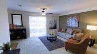 This single-story residence centers around a well-appointed living room that blends comfort and style. The space features neutral-toned carpeting underfoot, complemented by an accent wall painted in a muted olive green that adds depth and visual interest. A ceiling fan with a light fixture is centrally mounted, providing both air circulation and ambient lighting.

Large sliding glass doors allow generous natural light to fill the room while offering direct access to an outdoor balcony with views of surrounding greenery. The living area is furnished with a spacious gray sofa adorned with patterned throw pillows, paired with a warm-toned upholstered armchair, creating a cohesive and inviting seating arrangement. A dark-framed glass coffee table sits atop a textured area rug, anchoring the space.

Decorative wall art, including a typographic canvas and a framed photographic print, adds personality to the room. Crown molding along the ceiling provides a clean, finished look throughout. The overall ambiance is casual yet polished, making the living area a functional and aesthetically pleasing central hub of the home.