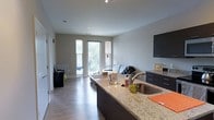This single-floor residence offers a thoughtfully designed open-concept layout that seamlessly connects the kitchen and living areas into one cohesive space. The kitchen is a standout feature, highlighted by a large granite-topped island with an undermount sink, dark cabinetry with ample storage, and contemporary finishes throughout. The countertops display attractive natural stone veining, lending both functionality and visual appeal to the space.

The living area flows naturally from the kitchen, benefiting from an abundance of natural light streaming through large windows and a glass-paneled door that opens onto a private balcony. The outdoor views of surrounding greenery further enhance the bright, airy ambiance of the interior. Light-toned, hardwood-style flooring runs throughout the space, adding warmth and continuity across both the kitchen and living zones.

The neutral gray walls and white ceilings complement the dark cabinetry and stone surfaces, creating a balanced, modern aesthetic. Recessed and track lighting provide flexible illumination, while the open layout ensures excellent sight lines throughout, making the space feel expansive and welcoming.
