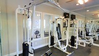 This single-floor fitness center offers a practical and well-equipped space designed to support strength training and general exercise needs. The room features a multi-station cable machine and a dedicated weight machine, both finished in white, providing a range of resistance training options. Dark rubber flooring runs throughout, offering durability and stability underfoot.

Mirrored walls line one side of the space, visually expanding the room and allowing users to monitor their form during workouts. Crown molding along the ceiling adds a refined architectural detail that elevates the overall finish of the space. Recessed lighting, combined with natural light filtering through interior windows, ensures the area remains bright and functional.

A wall-mounted television provides entertainment during workouts, adding to the convenience of the facility. Visible through the interior windows is an adjacent laundry area equipped with a washer and dryer, offering residents easy access to laundry facilities within the same building. The neutral wall tones and clean, organized layout contribute to a focused and efficient environment suited for everyday fitness use.