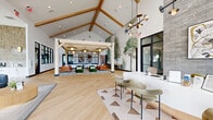 Located in Thornton, Colorado, this thoughtfully designed single-floor space spans nearly 2,000 square feet and serves as an inviting communal or leasing hub. The architecture is immediately striking, defined by soaring vaulted ceilings with exposed natural wood beam trusses that lend a warm, barn-inspired character to the otherwise contemporary interior. Light hardwood flooring laid in a chevron pattern flows throughout, unifying the open-concept layout.

At the heart of the space is a freestanding pergola-style structure with its own overhead beam framework, beneath which a curated lounge area features cognac leather sofas, plush emerald-green accent chairs, and woven pendant lighting — creating an intimate gathering spot within the larger room. Adjacent to this is a sleek white reception or workspace counter paired with velvet seating and anchored by a dramatic floor-to-ceiling gray brick accent wall adorned with artwork.

Nature-inspired murals of tall trees animate the white walls, reinforcing a biophilic design theme. Oversized windows along the perimeter flood the space with natural light and offer open views of the surrounding Colorado landscape. A hallway bordered by striking black-framed glass partitions connects to additional rooms, providing a sense of transparency and architectural interest. The overall ambiance is modern, airy, and welcoming.