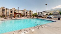 Located in South Ogden, Utah, this outdoor amenity area offers a well-appointed recreational space set against a dramatic backdrop of the Wasatch Mountains. The centerpiece is a generously sized swimming pool with clear, blue water, entry handrails, and clearly marked depth indicators, making it both inviting and functional. Surrounding the pool is a broad concrete deck furnished with multiple chaise lounges, providing ample space for sunbathing and relaxation.

A standout architectural feature is the stained wood pergola, which anchors a shaded seating and dining area complete with a round picnic table. The pergola's warm, rich tones complement the surrounding residential buildings, which feature a blend of lap siding, stone veneer accents, and pitched rooflines. The entire area is enclosed by a decorative wrought-iron fence with finial-topped posts, ensuring both security and an attractive perimeter.

A separate hot tub or spa is also present within the complex, adding to the recreational offerings. The open-sky setting allows for abundant natural light throughout the day, while the mountain views to the east create a scenic and tranquil atmosphere. The well-maintained hardscape, thoughtful landscaping, and cohesive design elements make this outdoor amenity space a key highlight of the property.