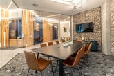 This thoughtfully designed single-floor space blends contemporary architecture with warm natural materials to create an inviting and functional environment. The expansive lobby serves as the centerpiece, featuring large floor-to-ceiling windows that flood the interior with natural light and offer views of the surrounding streetscape. Light-toned large-format tile flooring extends throughout, complementing the open, airy feel of the space. A built-in communal table with blue wire chairs and integrated planters adds a social focal point to the lobby area.

Natural wood-clad columns are a defining architectural element, providing warmth and texture that contrasts beautifully with the sleek glass partitions used to define separate zones within the space. Geometric linear LED lighting fixtures are suspended from the ceilings, adding a striking visual detail that reinforces the modern aesthetic.

A glass-enclosed conference room offers a semi-private meeting space furnished with a long dark wood table and cognac leather chairs. A rugged gabion stone accent wall anchors the room and provides a textural backdrop for the wall-mounted display screen. Adjacent areas visible through the glass partitions include a lounge and game space, enhancing the multi-functional nature of this well-appointed amenity floor.