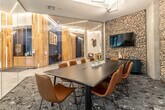This thoughtfully designed single-floor space blends contemporary architecture with warm natural materials to create an inviting and functional environment. The expansive lobby serves as the centerpiece, featuring large floor-to-ceiling windows that flood the interior with natural light and offer views of the surrounding streetscape. Light-toned large-format tile flooring extends throughout, complementing the open, airy feel of the space. A built-in communal table with blue wire chairs and integrated planters adds a social focal point to the lobby area.

Natural wood-clad columns are a defining architectural element, providing warmth and texture that contrasts beautifully with the sleek glass partitions used to define separate zones within the space. Geometric linear LED lighting fixtures are suspended from the ceilings, adding a striking visual detail that reinforces the modern aesthetic.

A glass-enclosed conference room offers a semi-private meeting space furnished with a long dark wood table and cognac leather chairs. A rugged gabion stone accent wall anchors the room and provides a textural backdrop for the wall-mounted display screen. Adjacent areas visible through the glass partitions include a lounge and game space, enhancing the multi-functional nature of this well-appointed amenity floor.
