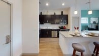 This well-appointed single-floor residence showcases a thoughtfully designed open-concept layout that seamlessly connects the kitchen, dining, and living areas. The kitchen is a standout, featuring dark espresso cabinetry contrasted by a white subway tile backsplash, sleek granite countertops, and a full suite of stainless steel appliances. A central island with pendant lighting above serves as both a prep space and casual dining spot, anchoring the room with style and function. Light wood-style flooring runs throughout the main living areas, lending warmth and continuity.

The living room makes a bold statement with a striking teal accent wall, offering a vibrant focal point against the neutral surroundings. The space is comfortable and well-lit, with a relaxed, modern aesthetic.

The primary bedroom is bright and inviting, highlighted by a vivid green accent wall, plush carpeting, and large windows that allow natural light to fill the room. A second bedroom provides a quiet, cozy retreat with direct sightlines to the kitchen area.

The bathroom is clean and modern, featuring a dark vanity with ample storage, a wide mirror with overhead lighting, and a full tub-shower combination lined with classic white subway tile. Every room reflects a cohesive, contemporary design sensibility throughout.