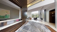 This striking lobby makes an immediate impression with its soaring double-height ceilings and expansive floor-to-ceiling curtain wall windows that flood the interior with natural light while framing lush outdoor greenery. The entry is defined by a bold combination of polished stone tile, rich hardwood flooring, and a large statement area rug in grey and gold geometric patterning, creating a dynamic visual foundation.

A prominent reception desk clad in horizontal dark wood slat paneling anchors one side of the lobby, complementing the warm tones found throughout the space. Opposite the entry, a sleek linear fireplace serves as a focal point, flanked by a decorative wall installation featuring an artful arrangement of circular gold and white elements. Contemporary lounge seating with colorful accent pillows is arranged thoughtfully nearby, encouraging conversation.

Stone-clad structural columns add architectural dimension and texture, while smooth white walls contrast elegantly with the darker wood and stone finishes. A corridor leads to additional spaces, including what appears to be a conference or meeting room visible through glass partitions. The overall ambiance is refined, modern, and welcoming, balancing professional sophistication with warm, curated design details.