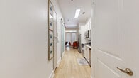 This single-floor studio is designed with both functionality and modern style in mind. Light wood-tone flooring runs throughout, creating a warm, cohesive feel that connects each area seamlessly. A long entry corridor leads into the heart of the unit, where the galley-style kitchen lines one side with crisp white shaker cabinets, a full suite of stainless steel appliances — including a side-by-side refrigerator with ice maker, electric range, over-range microwave, and dishwasher — and a clean white countertop with an undermount sink. The kitchen opens through a sleek sliding barn door into the combined living and dining area, which benefits from natural light filtering in through large windows with plantation shutters. The living area features notably tall ceilings that lend a sense of spaciousness, complemented by a bold accent wall. The bathroom is finished in all-white tile, featuring a full tub-shower combination, a vanity with white countertop, and overhead shelving for added storage. A dedicated closet provides practical organization. Throughout the space, white walls and recessed lighting maintain a bright, airy ambiance that feels open and inviting.