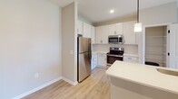 Located along Snowmass Loop in Garner, NC, this single-floor residence offers a clean, contemporary design throughout. The kitchen serves as a central highlight, featuring white shaker-style cabinetry, a herringbone tile backsplash, stainless steel appliances including a refrigerator, range, and over-the-range microwave, and a peninsula with light-toned countertops. Recessed lighting and a pendant fixture provide well-distributed illumination, while light wood-style flooring extends through the main living areas.

The living area is open and airy, anchored by a ceiling fan with light kit and French doors that lead to a private balcony, allowing natural light to flow in. Bedrooms are finished with plush carpet and ceiling fans; one bedroom features a wide window offering exterior views. Neutral wall tones carry consistently throughout the unit, contributing to a cohesive, modern aesthetic.

The bathroom is efficiently designed with a white vanity, matte black hardware accents, and matching wood-style flooring. A dedicated laundry room houses a full-size top-loading washer and dryer set with overhead wire shelving for additional storage. A pantry or utility closet is accessible directly from the kitchen area, adding functional convenience to the overall layout.