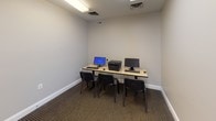 Located at 1201 Riverwalk Place in Frederick, Maryland, this single-floor space serves as a focused and functional business center. The layout is straightforward and efficiently designed to support productivity and shared use.

The primary area features a dedicated computer workstation room finished with neutral gray walls, clean white baseboards, and commercial-grade carpeting in a warm brown tone. A long workbench-style desk runs along one wall, accommodating multiple desktop computer monitors, keyboards, and a multifunction printer, with several task chairs positioned for easy access. Overhead fluorescent lighting ensures the room is well-lit, and ceiling-mounted HVAC vents provide climate control throughout.

The room is accessed through a classic six-panel white door, consistent with the clean, professional aesthetic of the space. A connecting hallway provides access to an adjacent tiled room visible in the background, which appears to serve as a utility or break area, equipped with a water cooler and waste receptacle. The transition between the carpeted and tiled zones is clearly defined, reinforcing the purposeful zoning of the space.

Overall, the space presents a clean, no-frills professional environment well-suited for shared computer access, administrative tasks, or small-scale office operations.