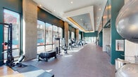 This expansive fitness center is designed with both functionality and aesthetics in mind, offering a premium workout environment. The space is defined by its striking deep teal accent walls, soaring ceilings, and an abundance of natural light streaming through large floor-to-ceiling windows that frame views of the surrounding urban streetscape. A coffered ceiling detail with warm ambient uplighting adds an upscale architectural touch to the main cardio area, complemented by multiple ceiling fans for airflow and comfort. The flooring transitions between speckled rubber and hardwood depending on the zone, providing appropriate surfaces for various workout styles. The cardio section is lined with a row of modern treadmills and elliptical machines, each equipped with individual digital screens. A dedicated strength and functional training zone features a power rack, cable machines, a battle rope, and a heavy tire for functional fitness. Spin bikes and rowing machines occupy a mirrored section of the space, with a large wall-mounted television for entertainment. A separate, open studio area with expansive glass walls and smooth flooring offers an ideal setting for stretching, yoga, or group fitness. The facility, branded as "The MAV," is thoughtfully laid out to accommodate a full spectrum of fitness activities.
