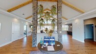 This single-floor community clubhouse is designed to impress from the moment you enter. The grand entrance is anchored by striking stacked stone columns that rise to meet soaring vaulted ceilings accented with warm, exposed wood beams. Cascading greenery suspended from the ceiling adds a natural, organic touch, while a central circular reception table serves as a welcoming focal point. Rich hardwood flooring runs throughout, tying the open layout together seamlessly.

The main lounge area offers comfortable seating arrangements beneath the dramatic cathedral ceiling, with ample natural light pouring in through large windows and expansive glass sliding doors. These doors open directly to an outdoor patio and pool area, creating a seamless indoor-outdoor connection. A wall-mounted flat-screen TV adds to the relaxed, social atmosphere.

The well-appointed kitchen features dark cabinetry, granite countertops, stainless steel appliances including a full refrigerator, microwave, and dishwasher, and a generous island with bar seating illuminated by elegant pendant lighting. The space is ideal for gatherings and community events.

Additional areas visible within the facility include what appears to be a fitness room, further enhancing the functionality of this thoughtfully designed community space.