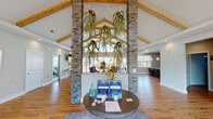 This single-floor community clubhouse is designed to impress from the moment you enter. The grand entrance is anchored by striking stacked stone columns that rise to meet soaring vaulted ceilings accented with warm, exposed wood beams. Cascading greenery suspended from the ceiling adds a natural, organic touch, while a central circular reception table serves as a welcoming focal point. Rich hardwood flooring runs throughout, tying the open layout together seamlessly.

The main lounge area offers comfortable seating arrangements beneath the dramatic cathedral ceiling, with ample natural light pouring in through large windows and expansive glass sliding doors. These doors open directly to an outdoor patio and pool area, creating a seamless indoor-outdoor connection. A wall-mounted flat-screen TV adds to the relaxed, social atmosphere.

The well-appointed kitchen features dark cabinetry, granite countertops, stainless steel appliances including a full refrigerator, microwave, and dishwasher, and a generous island with bar seating illuminated by elegant pendant lighting. The space is ideal for gatherings and community events.

Additional areas visible within the facility include what appears to be a fitness room, further enhancing the functionality of this thoughtfully designed community space.