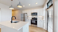 Located at 300 E Fifth Ave in Columbus, Ohio, this single-floor residence offers a thoughtfully designed open-concept layout with a clean, modern aesthetic. Light-toned hardwood flooring flows throughout the space, complementing the bright white walls and creating a cohesive, airy atmosphere.

The kitchen is a standout feature, showcasing white shaker-style cabinetry with a classic subway tile backsplash and quartz countertops. A generous center island with an undermount sink and matte black faucet serves as the focal point, illuminated by industrial-style pendant lights. Stainless steel appliances, including a refrigerator, range, and built-in microwave, complete the well-appointed space. Large floor-to-ceiling windows and a glass door offer views to an outdoor balcony, flooding the kitchen and living area with natural light.

A clean hallway with recessed lighting connects the living areas to the private spaces, including a well-proportioned bedroom with light wood flooring and a sliding barn door accent. The bathroom features a walk-in glass shower enclosed by matte black hardware and framing, paired with crisp vertical white tile — a striking contrast that adds a refined, contemporary touch. The overall design balances modern functionality with stylish details throughout.