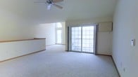 This single-floor space offers a welcoming and airy atmosphere, highlighted by vaulted ceilings that lend a sense of openness and volume to the main living area. A ceiling fan is centrally mounted overhead, providing both practical airflow and a classic finishing touch. Natural light floods the interior through multiple windows, including a charming arched window positioned high on the wall, as well as a large sliding glass door that opens to an outdoor balcony. The sliding door is framed by floor-length curtain panels and features a multi-pane grid design, offering expansive views of the exterior. Soft, plush wall-to-wall carpeting in a neutral tone covers the floor throughout, creating a comfortable and cohesive look. A partial interior wall or half-partition is visible toward the rear of the space, subtly defining areas within the open layout. The overall color palette is light and neutral, with white walls and warm wood-toned trim accents along the baseboards, contributing to a clean, move-in-ready aesthetic. The combination of vaulted ceilings, abundant natural light, and outdoor access makes this a bright and versatile living space.