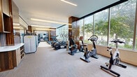 Located at 1575 Anderson Rd in McLean, Virginia, this professionally appointed fitness center offers a comprehensive workout environment within a generously sized single-floor space. The facility is designed with a modern aesthetic, incorporating rich wood-paneled accent columns, textured wall treatments, and sleek recessed and linear pendant lighting that contribute to a polished, upscale atmosphere.

A defining architectural feature is the expansive floor-to-ceiling window wall that runs along one side of the space, flooding the interior with abundant natural light and providing open views of the surrounding greenery. The neutral-toned carpet flooring and clean white ceilings further enhance the bright, airy ambiance.

The cardio zone is well-equipped with a row of treadmills, elliptical trainers, stationary bikes, and recumbent cycling machines, many featuring individual digital displays. A dedicated strength training area houses a comprehensive selection of Technogym selectorized machines, including leg press, chest press, and cable systems, as well as a dumbbell rack. Large wall-mounted televisions are positioned throughout for entertainment during workouts.

A glass-walled flex space provides an area suitable for stretching or functional training. Full-length mirrors enhance the sense of depth across the facility. The overall design balances functionality with refined aesthetics, making it a well-rounded fitness destination.