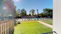 This outdoor amenity space centers around a generously sized swimming pool that serves as the focal point of the area. The pool is set within a well-maintained pool deck lined with numerous lounge chairs and seating, providing ample space for relaxation and recreation. A single-story clubhouse building runs along the back of the pool area, finished in a gray exterior with multiple windows that overlook the water. The structure appears to house support facilities for the pool area.

A secure perimeter fence encloses the entire pool and deck zone, ensuring both safety and privacy. Adjacent to the fenced pool area, a broad expanse of lush, well-kept green lawn opens up, offering additional outdoor space for leisure. A concrete walkway cuts through the grass, providing clear, direct access to the pool entrance.

The entire setting is framed by a backdrop of tall, mature deciduous trees that provide natural shade and a sense of seclusion. Under clear blue skies, the space benefits from abundant natural sunlight, creating a bright and inviting outdoor environment well-suited for warm-weather enjoyment.