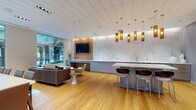 This thoughtfully designed single-floor space combines multiple functional zones within one open, flowing layout. Warm bamboo hardwood flooring runs throughout, providing a cohesive foundation that ties together the various areas seamlessly.

The lounge area is anchored by a generously sized sofa and contemporary accent chairs arranged around an organically shaped coffee table. Large floor-to-ceiling windows flood this zone with natural light and offer lush, tree-lined outdoor views, creating a relaxed yet refined atmosphere.

Adjacent to the lounge, a built-in media wall clad in warm wood paneling houses a flat-screen television, complemented by a low-profile credenza and shelving. A dramatic marble-look feature wall runs the length of the kitchen and bar area, serving as a striking backdrop for a sleek, extended counter finished in light stone with integrated bar seating below a cluster of amber glass pendant lights.

A dining zone with rectangular tables and upholstered chairs occupies the central-to-rear portion of the space, while a glass-partitioned room visible from the main area offers additional flexible use. Recessed and track lighting throughout ensures well-distributed illumination, while subtle wood ceiling accents add warmth and architectural detail to the overall refined, contemporary ambiance.