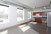 This single-floor urban loft showcases a compelling blend of industrial and contemporary design throughout its open layout. The living area is a standout feature, boasting tall ceilings with exposed ductwork and track lighting running along black pipe infrastructure — hallmarks of classic loft architecture. A row of large, floor-to-ceiling windows floods the space with abundant natural light and frames views of the surrounding cityscape, creating a bright and airy atmosphere.

The kitchen is well-appointed with warm medium-toned wood cabinetry, dark stone-finish countertops, and a full suite of stainless steel appliances, including a refrigerator with ice maker, electric range, built-in microwave, and dishwasher. A double stainless steel sink and ample upper and lower cabinet storage make this a highly functional cooking space. The kitchen's open orientation connects seamlessly to the living area.

The bedroom is a generously sized, quiet room finished with plush carpeting and clean white walls, offering a contrast to the more industrial common areas. An electrical panel is conveniently accessible within the room.

The bathroom features rich wood-toned cabinetry with brushed metal hardware, dark countertops, an undermount sink, and a full soaking tub with shower. Dark hardwood-style flooring ties together the modern aesthetic throughout the space.