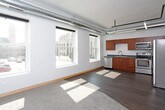 This single-floor urban loft showcases a compelling blend of industrial and contemporary design throughout its open layout. The living area is a standout feature, boasting tall ceilings with exposed ductwork and track lighting running along black pipe infrastructure — hallmarks of classic loft architecture. A row of large, floor-to-ceiling windows floods the space with abundant natural light and frames views of the surrounding cityscape, creating a bright and airy atmosphere.

The kitchen is well-appointed with warm medium-toned wood cabinetry, dark stone-finish countertops, and a full suite of stainless steel appliances, including a refrigerator with ice maker, electric range, built-in microwave, and dishwasher. A double stainless steel sink and ample upper and lower cabinet storage make this a highly functional cooking space. The kitchen's open orientation connects seamlessly to the living area.

The bedroom is a generously sized, quiet room finished with plush carpeting and clean white walls, offering a contrast to the more industrial common areas. An electrical panel is conveniently accessible within the room.

The bathroom features rich wood-toned cabinetry with brushed metal hardware, dark countertops, an undermount sink, and a full soaking tub with shower. Dark hardwood-style flooring ties together the modern aesthetic throughout the space.