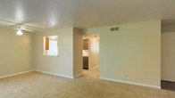 This single-story space features a spacious, open living area finished with wall-to-wall carpet in a warm, neutral tone. The walls are painted in a light beige, contributing to a bright and airy atmosphere throughout. A textured ceiling adds subtle visual interest overhead, while a ceiling-mounted HVAC vent ensures comfortable climate control. Natural light enters the living area through a window that also serves as a pass-through opening to the kitchen, offering a convenient connection between the two spaces. The kitchen is accessible through a dedicated doorway and is equipped with wooden cabinetry in a warm brown finish, providing ample storage. Appliances are visible within the kitchen, suggesting a functional and well-equipped cooking space. Clean white baseboards run along the perimeter of the room, providing a crisp contrast to the carpeted floors. The overall layout is straightforward and efficient, with an open flow between the living and kitchen areas. The neutral color palette and simple design elements create a clean, versatile environment suitable for a variety of interior arrangements and personal styles.