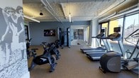 This fitness center offers a thoughtfully designed workout environment that combines industrial aesthetics with modern functionality. The space features an exposed ceiling with visible ductwork and piping, complemented by sleek linear lighting that provides bright, even illumination throughout. The walls are painted in a cool slate blue, creating a focused and energizing atmosphere.

The gym is outfitted with a comprehensive selection of equipment to support a variety of workout routines. Cardio options include multiple treadmills and Stages cycling bikes, catering to both high-intensity and endurance training. A cable machine and strength training station provide options for resistance workouts, while wall-mounted mirrors help maximize the sense of space and allow for form checks during exercise.

A mounted flat-screen television is positioned on the main wall, offering entertainment during workouts. The flooring consists of durable rubber-style gym matting, providing cushioning and stability underfoot. Large windows along one side of the room allow natural light to filter in, connecting the interior to the outdoors. A glass-partitioned area is visible toward the rear, suggesting additional designated zones within the facility. Overall, this fitness center delivers a clean, professional, and fully functional space for a wide range of athletic activities.