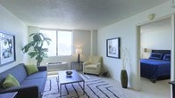 This single-floor space centers around a well-appointed living area that emphasizes natural light and a clean, understated aesthetic. The room is finished with light-colored walls and a textured ceiling, contributing to a calm and airy atmosphere. A large window dominates one wall, allowing generous amounts of natural light to fill the space while offering outward views, enhancing the sense of openness. The flooring is carpeted in a neutral tone, complemented by a patterned area rug that anchors the seating arrangement. The living area accommodates a sofa and an armchair, arranged around a low-profile coffee table to create an inviting, functional lounge space. A wall-mounted framed artwork adds a decorative focal point, while a tall floor vase with dried branches introduces a natural, sculptural element. A side table with a table lamp provides warm supplemental lighting. A wall-mounted HVAC unit is positioned beneath the window, offering climate control. The overall ambiance is neutral and versatile, presenting a move-in-ready space with a straightforward, practical design sensibility suited for a range of living arrangements.