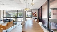This professional conference room is a compact yet thoughtfully designed meeting space that delivers a polished, modern aesthetic. The room is defined by its striking floor-to-ceiling glass wall and partition system, framed in dark metal, which allows natural light to flow freely throughout while maintaining a sense of privacy. Large exterior windows further enhance the bright, open atmosphere, offering views of surrounding greenery.

At the heart of the room sits a sleek, elongated wood conference table with built-in power and data ports, surrounded by clean-lined white upholstered chairs that seat a comfortable number of attendees. Light wood-toned flooring adds warmth and continuity to the space, complementing the neutral wall tones.

A wood credenza along one wall provides functional storage and surface space, accompanied by a multifunction printer and a decorative orchid arrangement. A bold, colorful abstract artwork anchors the wall above, adding a creative focal point to the otherwise refined interior. Cylindrical drum pendant lights hang from the drop-tile ceiling, casting a warm, even glow across the table.

The glass partitions open directly to a larger lobby or common area featuring dramatic green marble columns and lounge seating, extending the professional and upscale character of the overall environment.