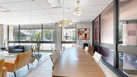 This professional conference room is a compact yet thoughtfully designed meeting space that delivers a polished, modern aesthetic. The room is defined by its striking floor-to-ceiling glass wall and partition system, framed in dark metal, which allows natural light to flow freely throughout while maintaining a sense of privacy. Large exterior windows further enhance the bright, open atmosphere, offering views of surrounding greenery.

At the heart of the room sits a sleek, elongated wood conference table with built-in power and data ports, surrounded by clean-lined white upholstered chairs that seat a comfortable number of attendees. Light wood-toned flooring adds warmth and continuity to the space, complementing the neutral wall tones.

A wood credenza along one wall provides functional storage and surface space, accompanied by a multifunction printer and a decorative orchid arrangement. A bold, colorful abstract artwork anchors the wall above, adding a creative focal point to the otherwise refined interior. Cylindrical drum pendant lights hang from the drop-tile ceiling, casting a warm, even glow across the table.

The glass partitions open directly to a larger lobby or common area featuring dramatic green marble columns and lounge seating, extending the professional and upscale character of the overall environment.