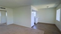 This single-floor space showcases a clean, minimal design with an open layout that connects the living area to the kitchen zone. The living area is finished with wall-to-wall carpet in a neutral beige tone, creating a soft and comfortable underfoot feel. A partial wall divides the living space from the kitchen area, providing a sense of separation while maintaining an open flow between the two zones. The kitchen features hard surface flooring, contrasting with the carpeted living area, and includes white cabinetry and a built-in countertop or breakfast bar along the perimeter. A window dressed with white vertical blinds allows natural light to filter into the kitchen, brightening the space. The walls throughout are painted in a clean, crisp white, contributing to a bright and airy atmosphere. Ceilings feature a subtle textured finish and are consistent in height throughout. The overall design is straightforward and functional, offering a neutral palette that provides flexibility for a variety of furnishing styles. The transition between the carpeted and hard-floor areas adds visual definition between the living and kitchen spaces.