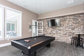 Located at 125 N Mill Rd in Vineyard, Utah, this dedicated game room is thoughtfully designed for leisure and social entertainment. The space is anchored by a full-size billiards table with a sleek gray felt surface, positioned beneath a modern linear pendant light that provides focused illumination for gameplay. One of the room's most striking architectural features is a floor-to-ceiling stacked stone accent wall, which lends a sophisticated, lodge-inspired aesthetic and serves as the backdrop for a wall-mounted flat-screen television. Opposite the stone wall, warm taupe walls are adorned with a curated gallery of black-and-white framed artwork, adding a refined, gallery-like character to the space. Large windows with plantation shutters line two walls, flooding the room with natural light and offering views of the surrounding community. High ceilings enhance the open, airy feel throughout. Neutral patterned carpet covers the floor, contributing to a comfortable and inviting atmosphere. A tall bistro table with bar stools is positioned near the windows, providing an ideal spot for spectators or casual conversation. Cue racks are mounted on the wall, keeping the space organized and ready for play. Overall, the room combines functional amenities with polished design details.