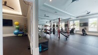 This spacious fitness center is thoughtfully designed to support a wide range of workout styles and training goals. The facility is organized into distinct zones, each outfitted with professional-grade equipment. The cardio area features multiple treadmills, elliptical machines, a stair climber, and stationary bikes, all positioned near large windows fitted with plantation shutters that admit generous natural light and offer views of lush greenery outside. A wall-mounted flat-screen television serves this section for entertainment during cardio sessions.

The strength training area is anchored by a bold red functional training rig equipped with a TKO punching bag, pull-up bars, and various cable attachments. An extensive free weight section includes a full rack of kettlebells and dumbbells alongside adjustable benches and a cable machine. Full-length mirrors line portions of the walls, enhancing the sense of openness and aiding in form correction.

Architecturally, the space features tray ceilings with crown molding, recessed lighting, and ceiling fans throughout, contributing to a polished yet functional aesthetic. Durable rubber flooring covers the entire workout area, providing safety and comfort. A dedicated entryway connects to adjacent storage and accessory areas, with medicine balls and foam rollers readily accessible.