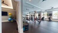 This spacious fitness center is thoughtfully designed to support a wide range of workout styles and training goals. The facility is organized into distinct zones, each outfitted with professional-grade equipment. The cardio area features multiple treadmills, elliptical machines, a stair climber, and stationary bikes, all positioned near large windows fitted with plantation shutters that admit generous natural light and offer views of lush greenery outside. A wall-mounted flat-screen television serves this section for entertainment during cardio sessions.

The strength training area is anchored by a bold red functional training rig equipped with a TKO punching bag, pull-up bars, and various cable attachments. An extensive free weight section includes a full rack of kettlebells and dumbbells alongside adjustable benches and a cable machine. Full-length mirrors line portions of the walls, enhancing the sense of openness and aiding in form correction.

Architecturally, the space features tray ceilings with crown molding, recessed lighting, and ceiling fans throughout, contributing to a polished yet functional aesthetic. Durable rubber flooring covers the entire workout area, providing safety and comfort. A dedicated entryway connects to adjacent storage and accessory areas, with medicine balls and foam rollers readily accessible.